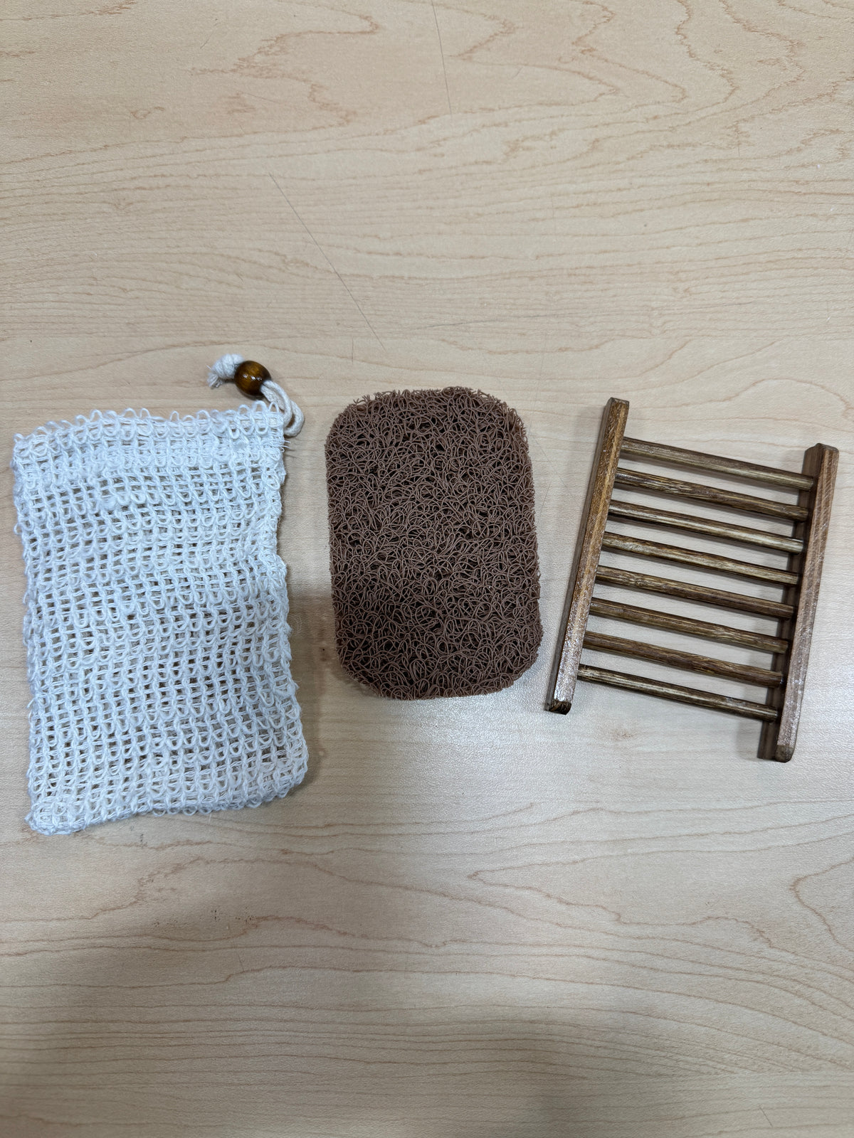 Three miniature bath accessory items arranged with a white background: a white crocheted exfoliating soap bag with a drawstring, a brown rectangular soap saver, and a small wooden soap dish.